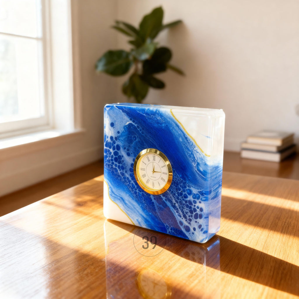 A square clock with a gold-colored face and Roman numerals. The clock's body features a blue and white marbled design with gold accents. The clock is sitting on a wooden surface.