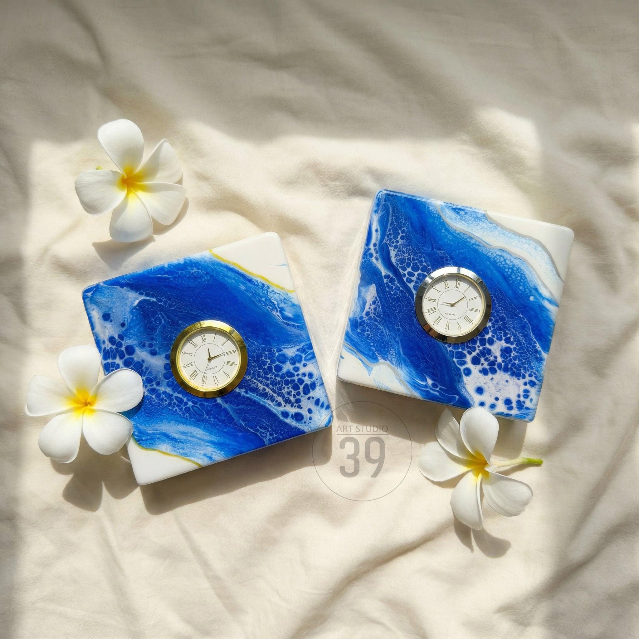 Two square clocks with a blue and white abstract design, each featuring a gold-colored clock face. The clocks are decorated with white and yellow flowers. The clocks are on a white surface.