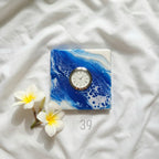 Square clock with a white face and silver bezel set into a blue and white abstract design. The clock face has black Roman numerals. The words "ART STUDIO 39" are printed on the bottom right.
