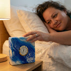 Square alarm clock with a blue and white abstract design. The clock face is gold with black numerals and hands. The clock is on a wooden nightstand next to a lamp.