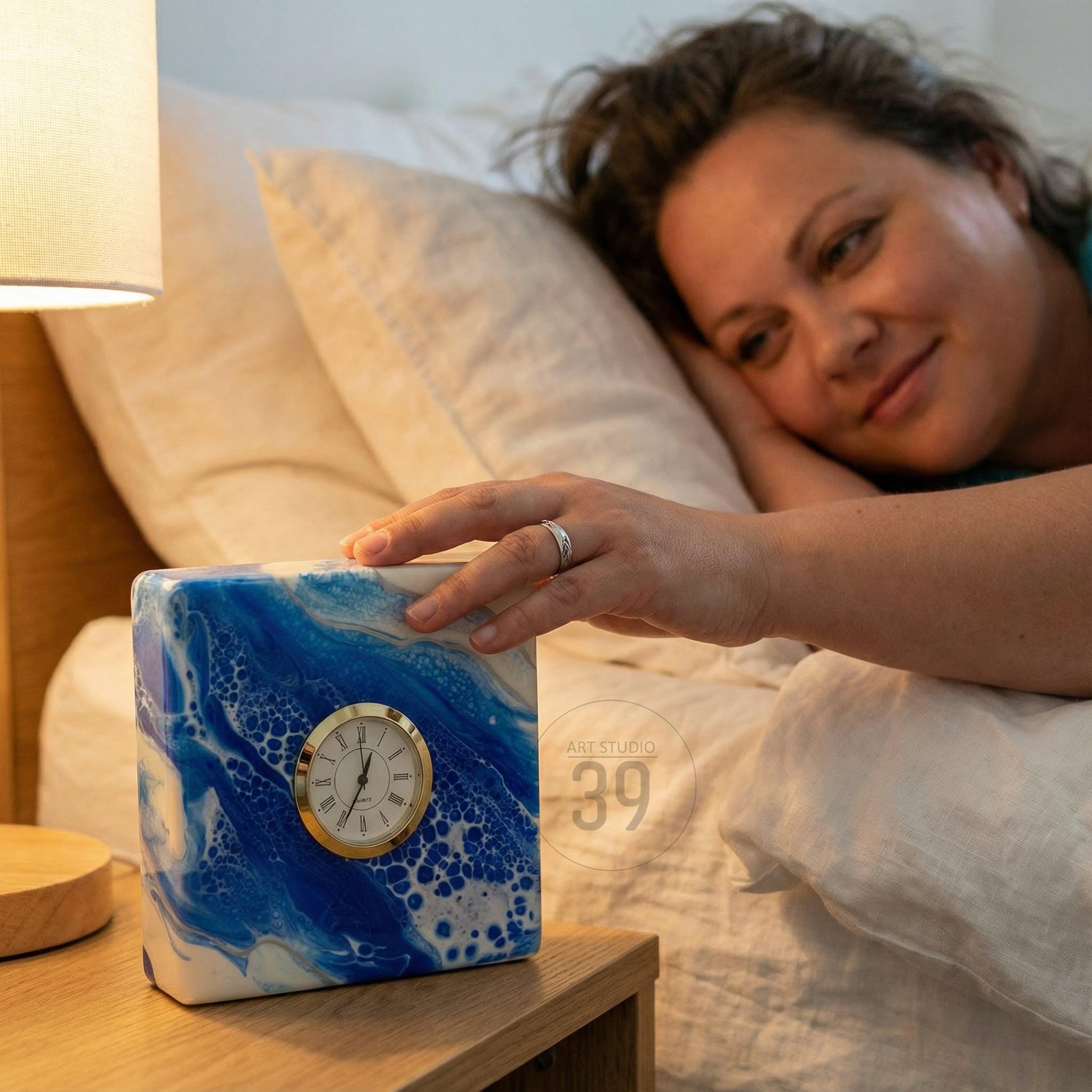 Square alarm clock with a blue and white abstract design. The clock face is gold with black numerals and hands. The clock is on a wooden nightstand next to a lamp.