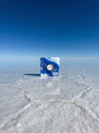 A square clock with a white face and silver hands is set into a cube with a blue and white marbled design. The clock is reflected in the water below. The number "39" is visible in the background.
