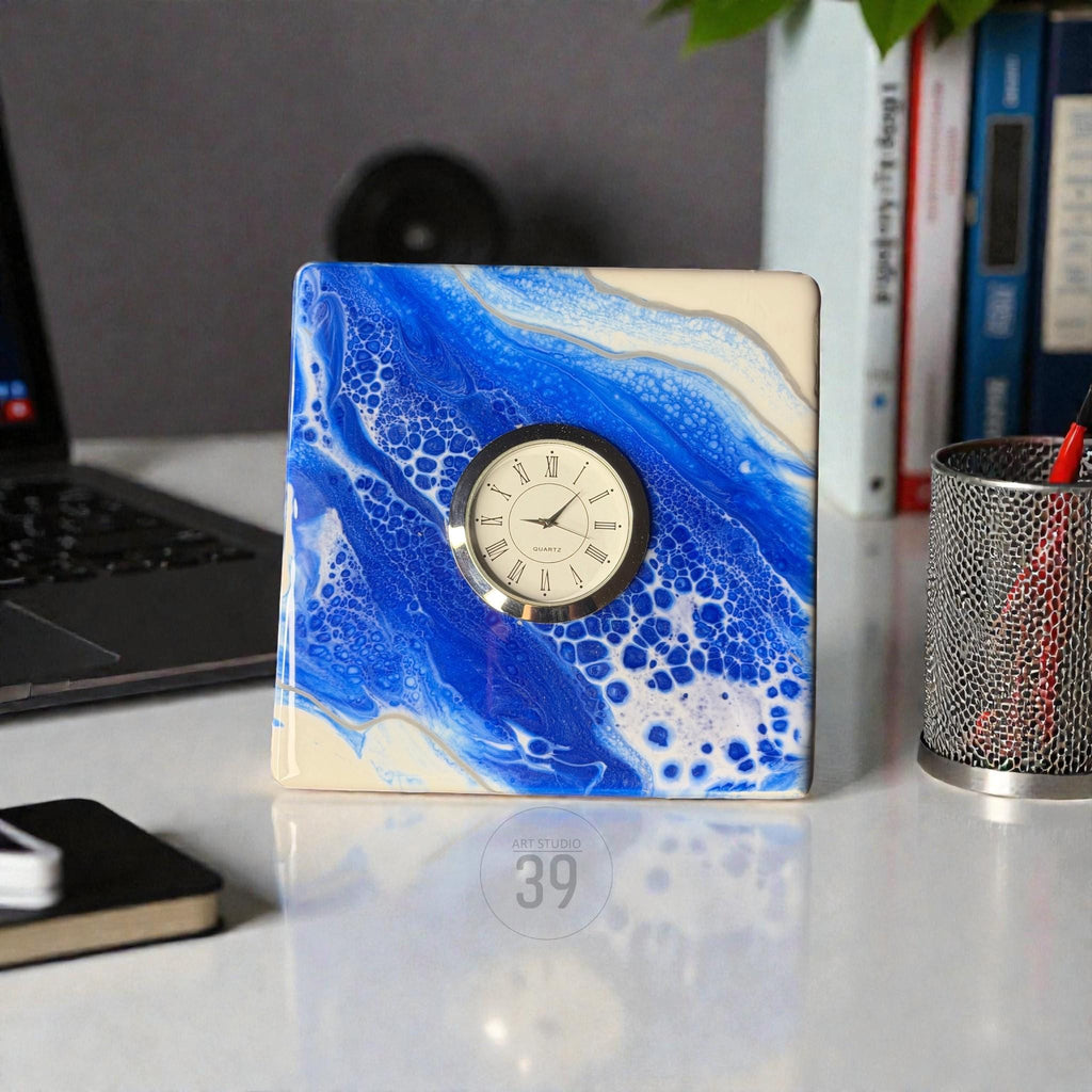 Square desk clock with a blue and white abstract design, featuring a silver-tone clock face with black Roman numerals. The clock has a quartz movement and is set on a white surface. The text "39" is visible on the surface.