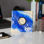 Square desk clock with a blue and white abstract design, featuring a silver-tone clock face with black Roman numerals. The clock has a quartz movement and is set on a white surface. The text "39" is visible on the surface.