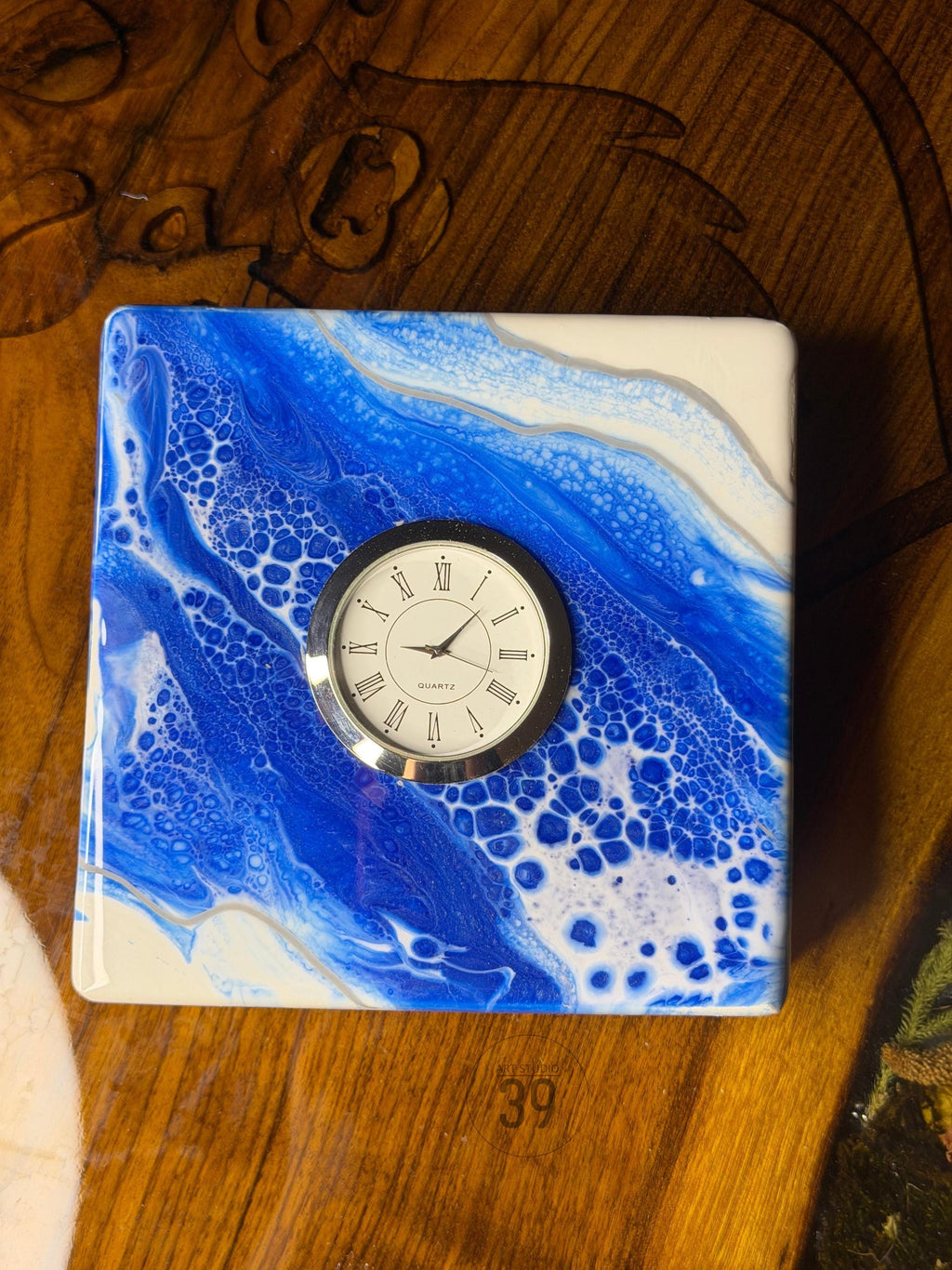 Square decorative clock with a blue and white abstract design. The clock face is round with Roman numerals and a silver bezel. The clock is labeled "Quartz".