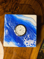 Square decorative clock with a blue and white abstract design. The clock face is round with Roman numerals and a silver bezel. The clock is labeled "Quartz".