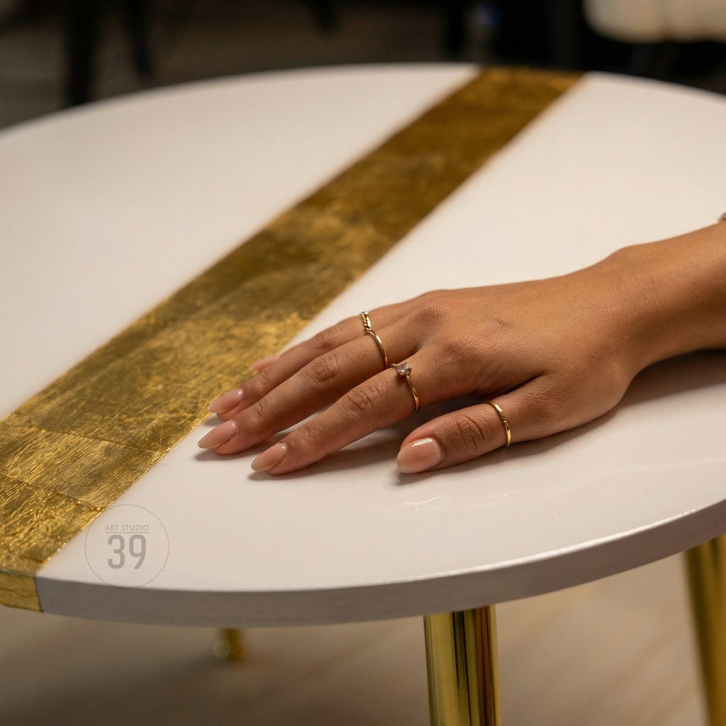 A white round table with a gold stripe down the center. The table has gold legs and a hand with several gold rings on it. The table has the text "ART STUDIO 39" on the side.