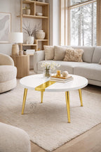 A modern, round coffee table with a white top and gold accents. The table features a gold stripe down the center and gold legs. The table is surrounded by neutral-toned furniture and decor, creating a contemporary living room setting.