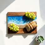 Wooden serving board with a resin ocean design. The board features a dark wood frame with a blue and white resin center, resembling ocean waves. Green grapes and sliced bread are arranged on the board, with a small succulent plant in a pot.