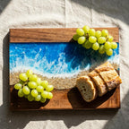A rectangular wooden serving board with a resin ocean design. The board features a blue and white wave pattern, a sandy beach area, and a dark wood border. Green grapes and sliced bread are arranged on the board. The number "39" is etched on the wood.
