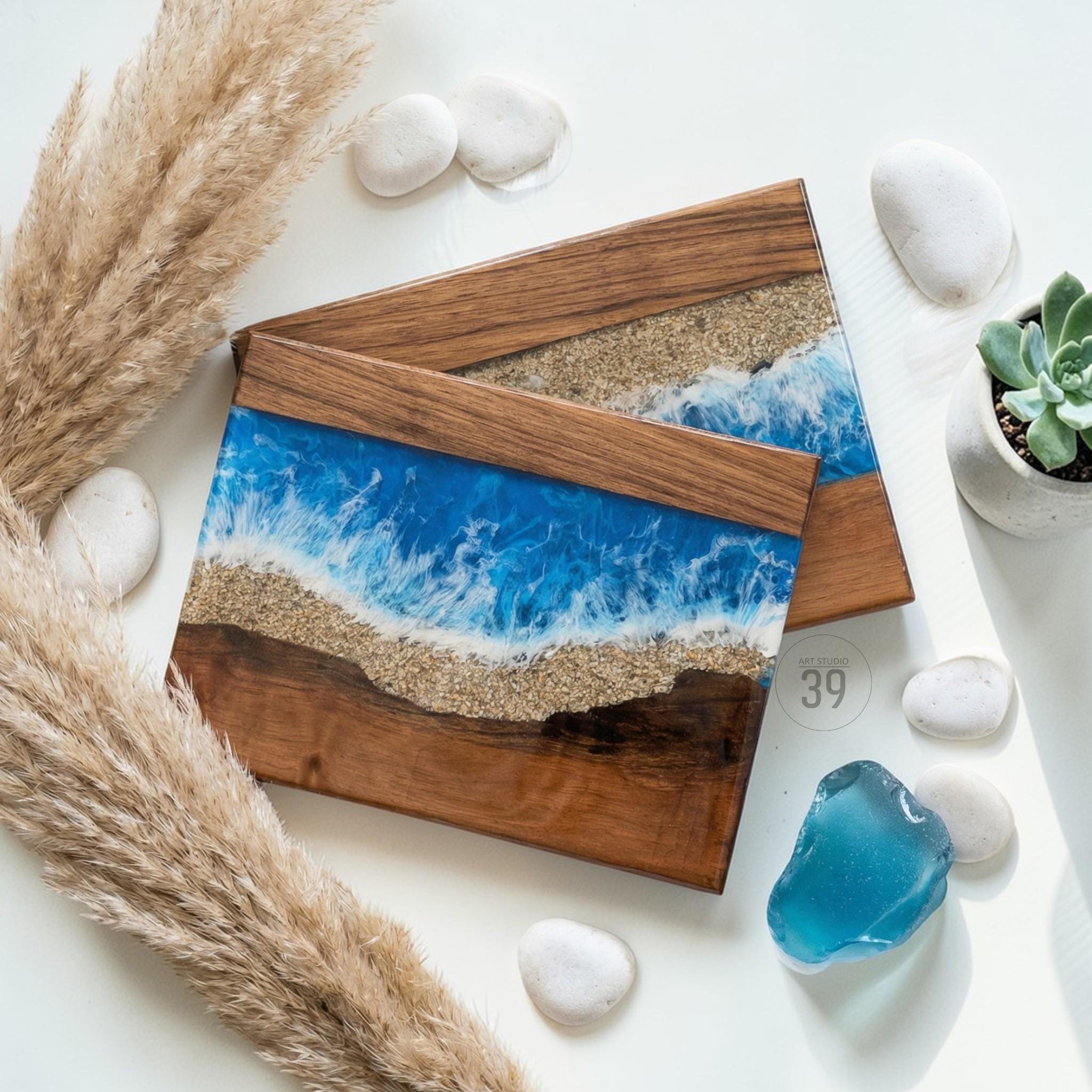Two wooden serving boards with a resin ocean design. The boards have a dark wood frame, a sandy beach layer, and a vibrant blue ocean with white waves. Decorative elements include white stones, a succulent, and dried grass.