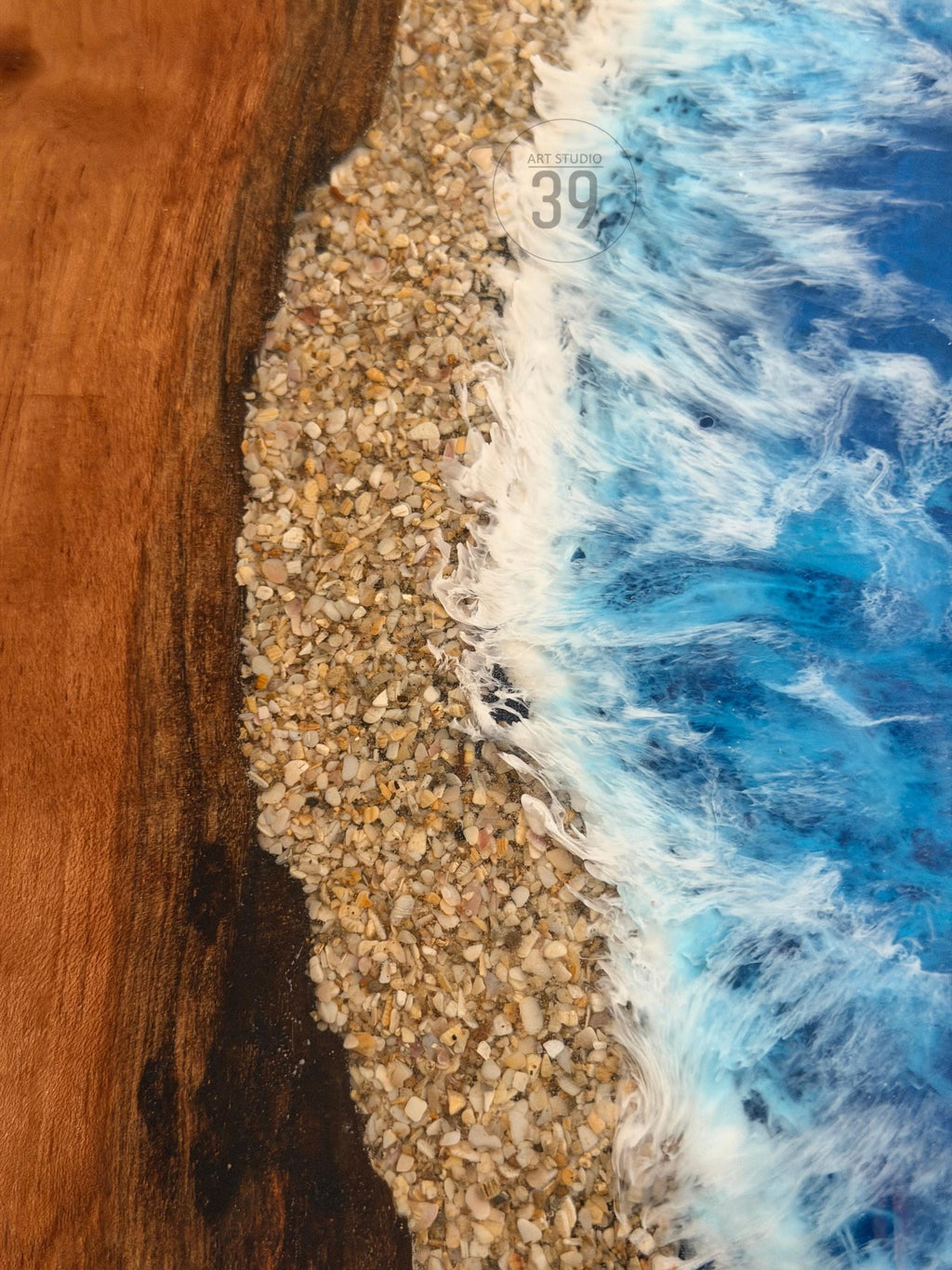 Close-up of a wooden surface with a beach scene. The design features a sandy shore made of small pebbles, with foamy white waves and blue water. The text "ART STUDIO 39" is visible.