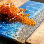 A decorative resin serving board with a beach scene and orange caviar. The board features a blue ocean design with white waves and a sandy shore. The caviar is garnished with small flowers and salt.