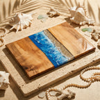 A rectangular wooden serving board with a blue resin wave design and a sandy beach section. The board is surrounded by seashells, pearls, and coral, creating a coastal theme. The number "39" is visible on the board.