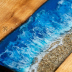 Wooden table with a blue resin river design, resembling ocean waves. The resin is a vibrant blue with white foamy details, and the edge of the table has a sandy beach effect. The number "39" is visible on the wooden surface.