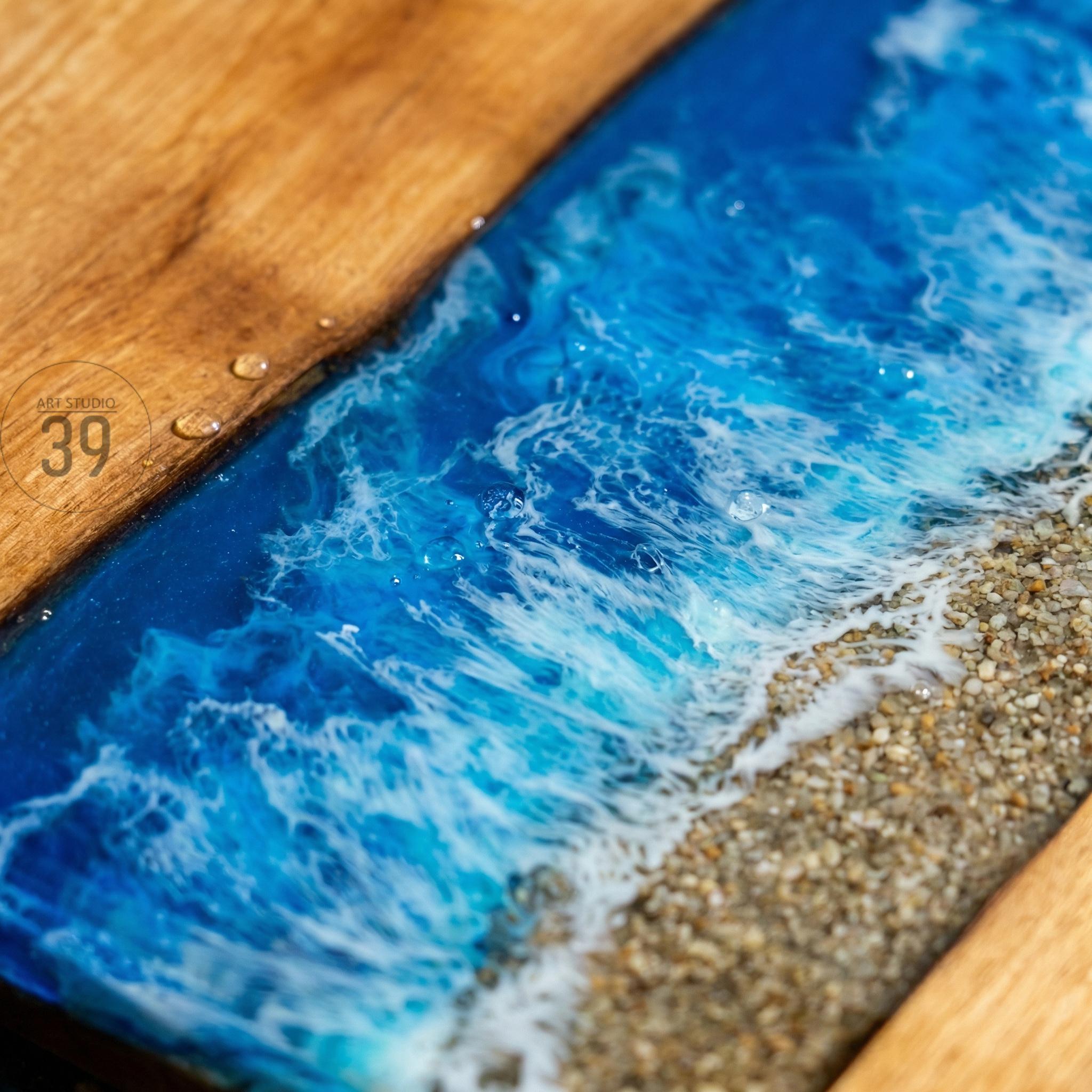 Wooden table with a blue resin river design, resembling ocean waves. The resin is a vibrant blue with white foamy details, and the edge of the table has a sandy beach effect. The number "39" is visible on the wooden surface.