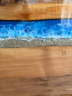 A wooden table with a blue resin river design, resembling ocean waves. The design includes a sandy beach effect and the text "Art Studio 39". The wood has a natural grain.