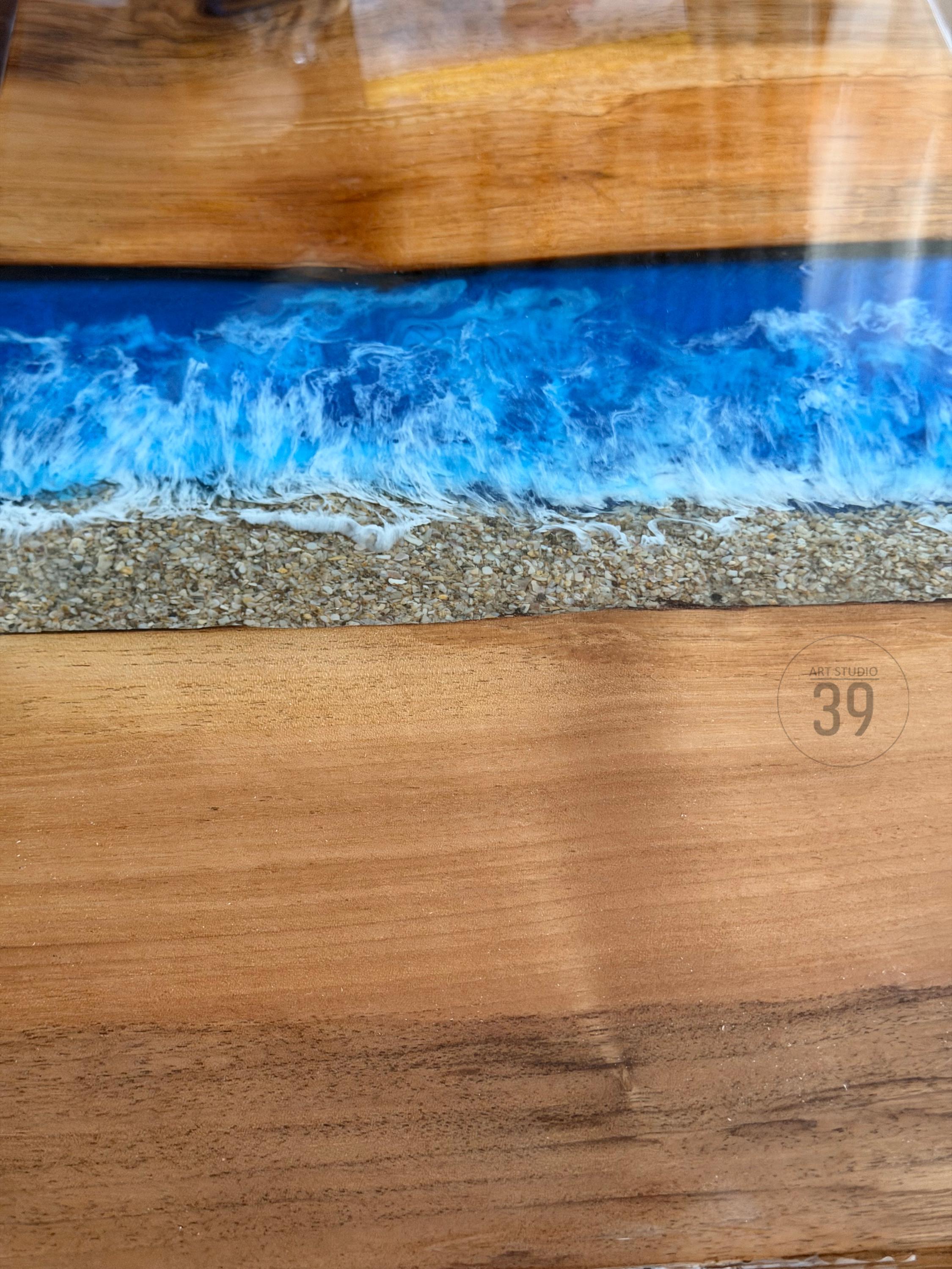 A wooden table with a blue resin river design, resembling ocean waves. The design includes a sandy beach effect and the text "Art Studio 39". The wood has a natural grain.