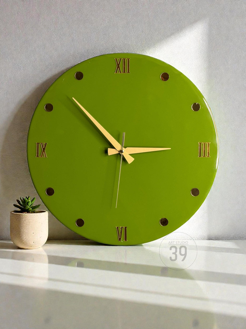 A round, lime green wall clock with gold-colored hands and Roman numeral hour markers. The clock has a minimalist design and is set against a white background. A small potted succulent is to the left of the clock.