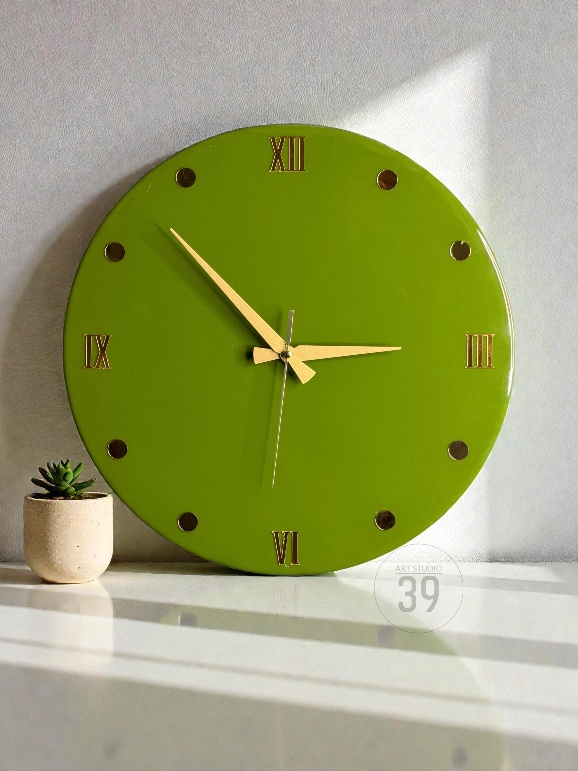 A round, lime green wall clock with gold-colored hands and Roman numeral hour markers. The clock has a minimalist design and is set against a white background. A small potted succulent is to the left of the clock.