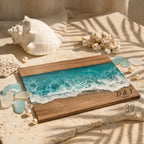 Wooden serving board with a resin ocean design. The board features a turquoise and white wave pattern, with a sandy beach effect. The initials "D &amp; Y" and the date "17.05.2023" are engraved on the board. The board is surrounded by seashells and pearls.