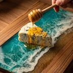 A wooden serving board with a resin ocean design, featuring a slice of blue cheese being drizzled with honey. The board has a wooden frame and a sandy beach effect.