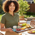 A woman in a green polo shirt holds a wooden serving board with various cheeses, meats, grapes, and bread. The board has a blue and white design on one end and the number 39. The background shows an outdoor setting.