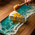 A wooden serving board with a blue and white resin design resembling ocean waves. A slice of blue cheese is drizzled with honey from a wooden honey dipper. The board has a wooden frame.