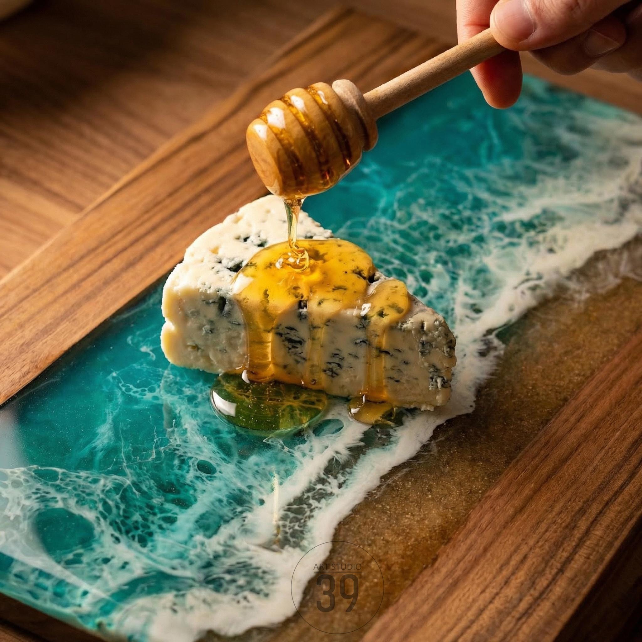 A wooden serving board with a blue and white resin design resembling ocean waves. A slice of blue cheese is drizzled with honey from a wooden honey dipper. The board has a wooden frame.