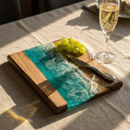 Wooden serving board with a turquoise resin design resembling ocean waves. The board has a natural wood border and a stainless steel knife. A bunch of green grapes rests on the board.