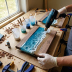 A woodworking project in progress, featuring wooden boards with a resin ocean design. The boards have a dark wood frame, with a blue and white resin ocean scene, and a sandy beach effect. A heat gun is being used on the resin.