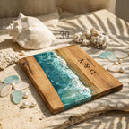 Wooden cutting board with a resin ocean design. The board features a wave pattern in shades of blue and white, with wooden sections on either side. The initials "D &amp; Y" and the date "17.05.2023" are engraved on the wood.