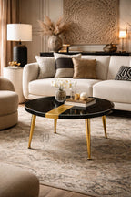 A round black coffee table with gold accents and legs. The table has a gold stripe down the center and is set on a patterned rug. Decorative pillows and a white sofa are in the background.