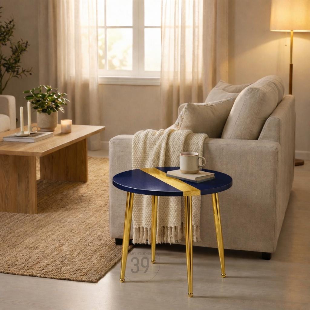 A navy blue and gold accent table with a round top and three gold legs. The table has a gold stripe across the top. A mug and a book sit on the table. The table is in a living room setting.