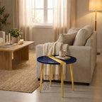 A navy blue and gold accent table with a round top and three gold legs. The table has a gold stripe across the top. A mug and a book sit on the table. The table is in a living room setting.