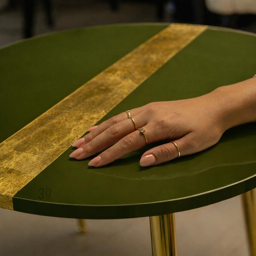 A round, green side table with a gold stripe and gold legs. The table has a glossy finish. A hand with gold rings rests on the table. The number "39" is visible on the table.