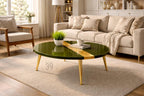 A round, green coffee table with gold-colored legs and a gold stripe down the center. A small plant, a book, and a white mug sit on the table. The table is in a living room with a beige rug and sofa.