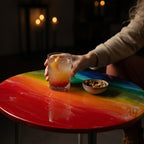 A round table with a vibrant rainbow-colored epoxy resin top. A clear glass with a cocktail and a small bowl of nuts sit on the table. The table has a glossy finish and a metal base.