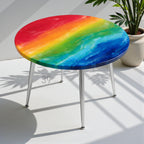 A round table with a rainbow-colored top and silver legs. The tabletop features a vibrant blend of red, orange, yellow, green, blue, and indigo hues. The table has three slender, silver legs.