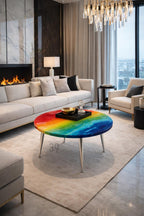 A modern living room features a rainbow-colored coffee table with silver legs. The table's surface displays a vibrant spectrum of colors, from red to blue, resembling an abstract painting. A black tray with a gold bowl and glass decor sits on top. The text "ART STUDIO 39" is visible.