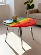 A round coffee table with a rainbow-colored top. The table has a glossy finish and chrome legs. A beige mug and a small book rest on the table. The text "ART STUDIO 39" is printed on the table.