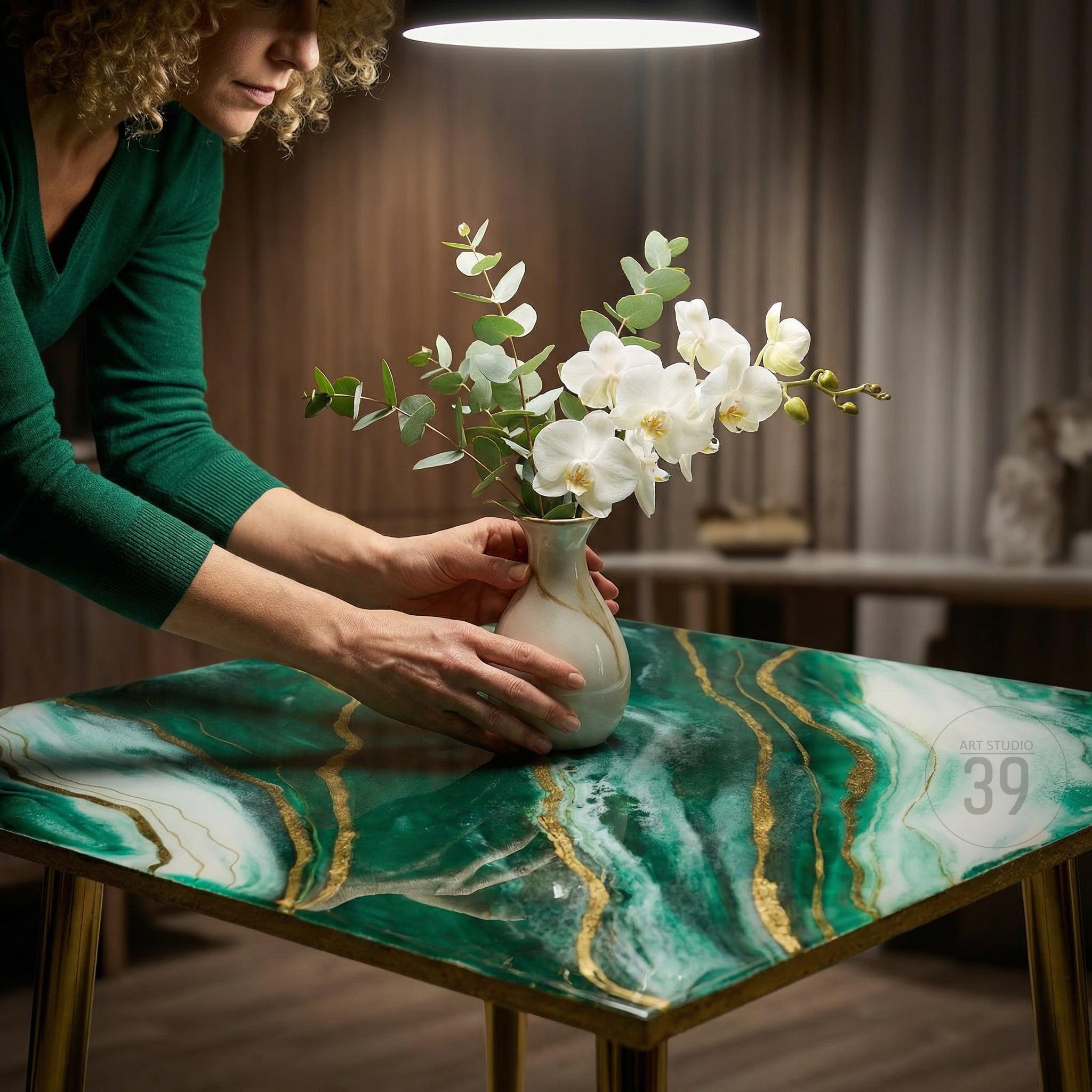 A square table with a green and gold marbled epoxy resin top. The table has gold metal legs. A white vase with white orchids and green eucalyptus is on the table. The table is in a well-lit room.