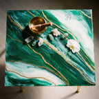 A square side table with a green and white marbled epoxy resin top, accented with gold veins. A brass singing bowl, crystals, and a white orchid are arranged on the surface. The table has gold-colored legs and the text "ART STUDIO 39".