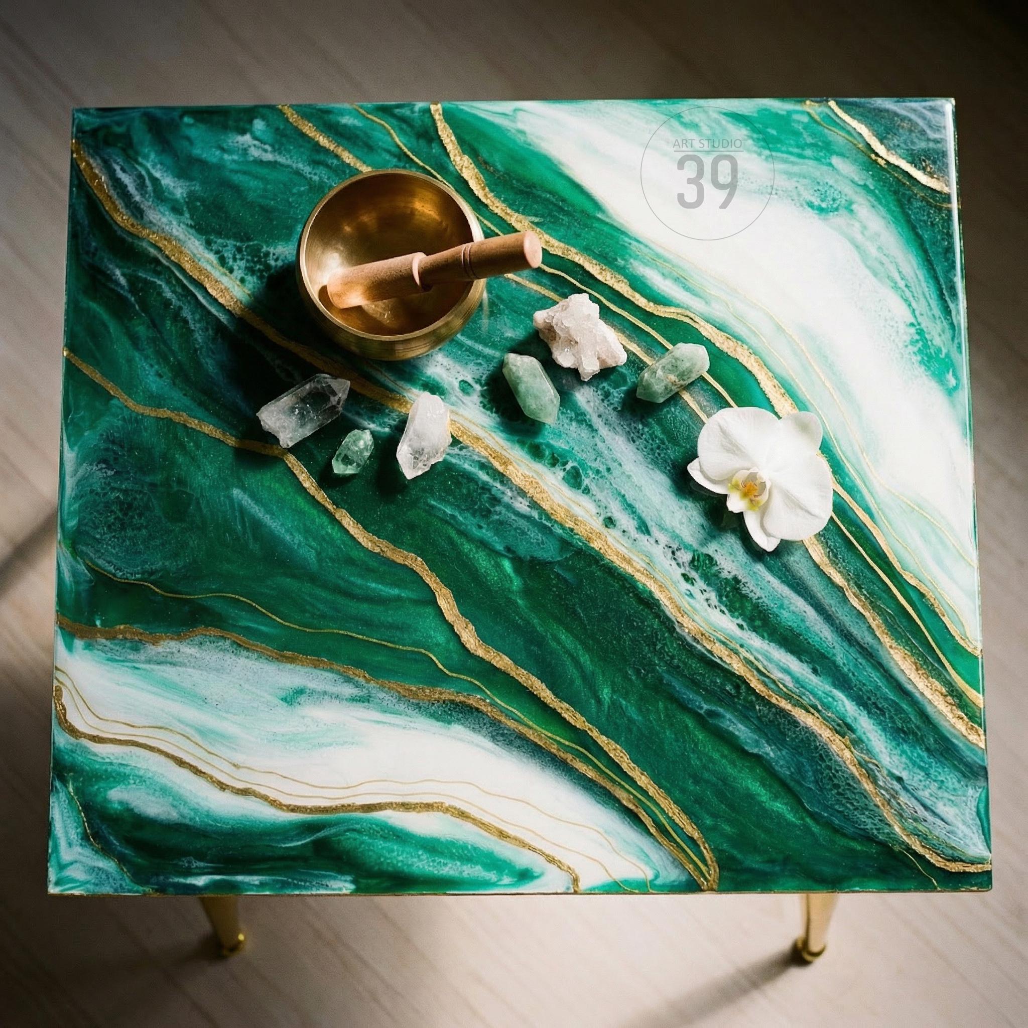 A square side table with a green and white marbled epoxy resin top, accented with gold veins. A brass singing bowl, crystals, and a white orchid are arranged on the surface. The table has gold-colored legs and the text "ART STUDIO 39".
