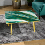 A square coffee table with a green and white marble-like top and gold-colored legs. The tabletop features gold-colored veining. The table is on a gray shag rug.