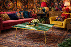 A living room scene featuring a red sofa with patterned pillows, a yellow armchair, and a green and gold coffee table. The room has floral wallpaper, a patterned rug, and decorative lamps.