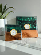Two square resin clocks with gold-tone clock faces. The clocks have a wooden base and a top layer of green and black resin. The smaller clock is on the left, and the larger clock is on the right. The clocks are on a white surface.