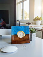 A square desk clock with a gold-colored bezel and a white clock face with black Roman numerals. The clock is set in a wooden base with a blue resin top. The clock is on a white desk with a computer mouse and a succulent plant.