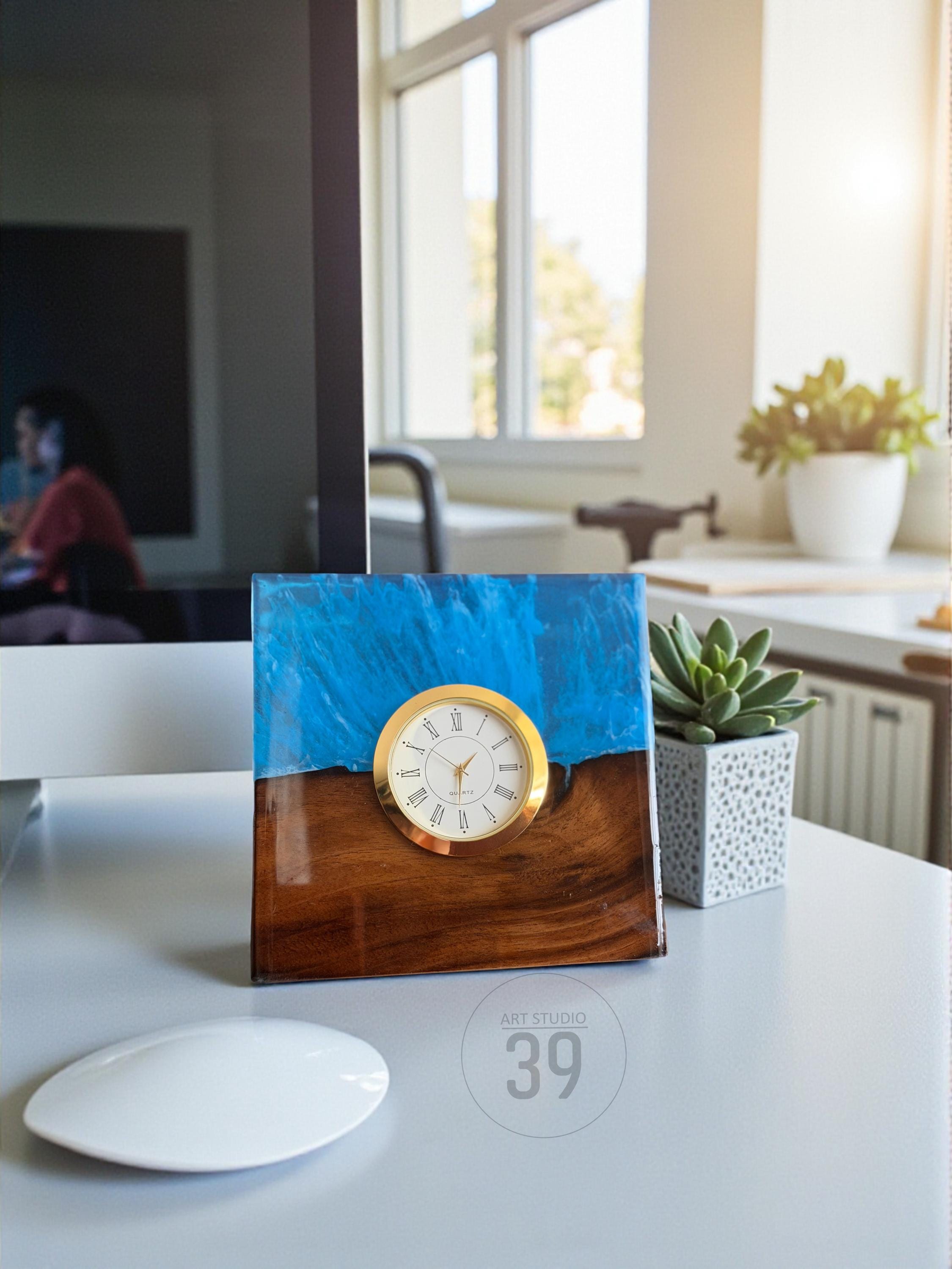 A square desk clock with a gold-colored bezel and a white clock face with black Roman numerals. The clock is set in a wooden base with a blue resin top. The clock is on a white desk with a computer mouse and a succulent plant.