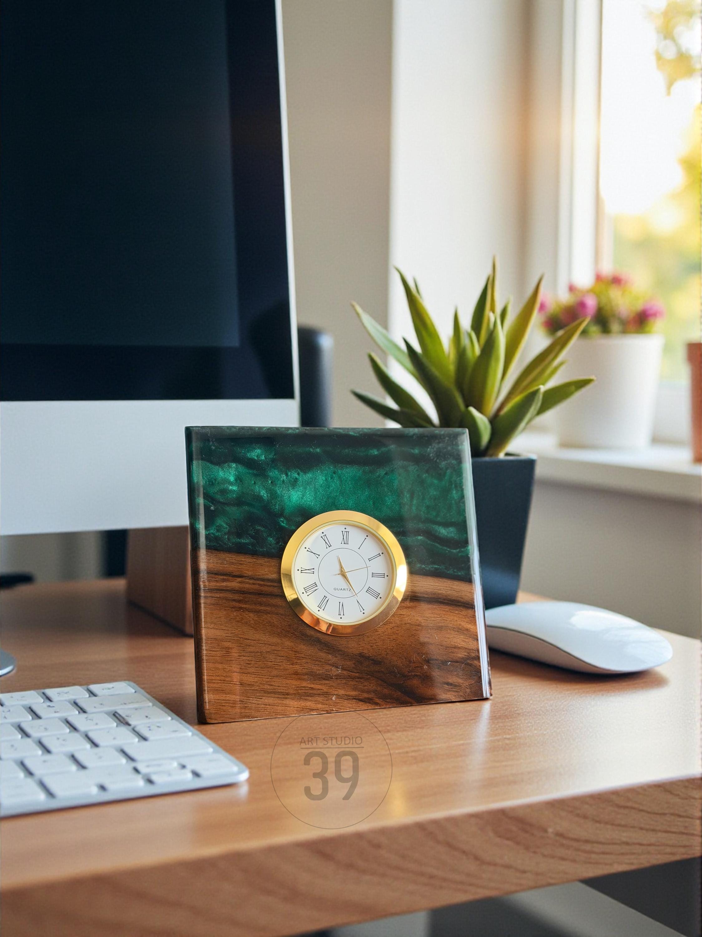 A square desk clock with a gold-colored bezel and white clock face. The clock is set in a wooden base with a green and blue resin top. The clock is on a wooden desk with a computer, keyboard, and mouse.